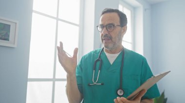 İspanyol doktor hastane odasında pano ve steteskopla el kol hareketi yapıyor.