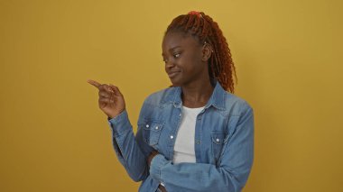 Kot giymiş genç bir Afrikalı Amerikalı kadın, sarı izole bir arka planı işaret ediyor.