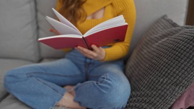 Oturma odasında kitap okuyan genç bir kadın. Kanepede dinlenirken sarı bir kazak ve kot pantolon giyiyor..