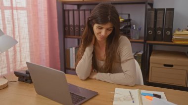 Ev ofisinde çalışan genç bir İspanyol kadın, bir dizüstü bilgisayar ve çeşitli ofis malzemeleriyle bir masada oturuyor, düşünceli ve odaklanmış görünüyor..