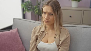 Güzel, genç, sarışın bir kadın, dairesinin oturma odasındaki bir kanepede düşünceli bir şekilde oturur. Huzurlu ve iç mekan manzarası yaratır..