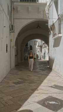 Genç İspanyol kadın güneşli bir günde Puglia, İtalya 'da bir kemer yolunun altında yaşlı bir kasaba caddesinde yürüyor..