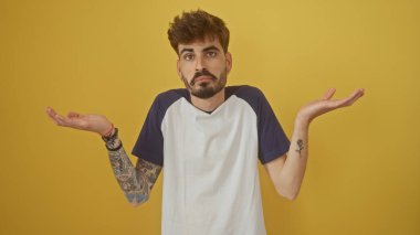 A confused young hispanic man with a beard and tattoos stands shrugging against a solid yellow background clipart