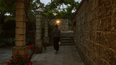 Bir kadın taştan duvarlar, canlı çiçekler ve bir pergola altında loş ışıklar ile çevrili, İtalya 'da kırsal bir puglia içinde bir patika boyunca yürür..