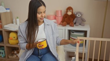 Kadın, oyuncaklar ve sakin dekorla çevrili bir beşiğe nazikçe dokunurken, davetkar bir yatak odasında kahveden zevk alıyor, anneliği ve sükuneti simgeliyor..