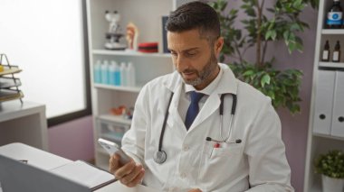 Klinikte tıbbi ekipman ve bitkilerle akıllı telefon kullanan beyaz önlüklü doktor hastane ortamındaki teknolojiye odaklanmış bir erkek sağlık uzmanı..