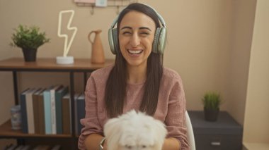 Kulaklıklı, gülümseyen genç bir kadın sıcak bir oturma odasında köpeğiyle vakit geçiriyor..