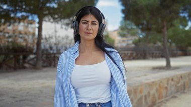 Genç, esmer, kulaklık takan, açık hava parkında ağaçlar ve binalarla çevrili mavi çizgili tişörtlü bir kadın kameraya güvenle bakıyor..