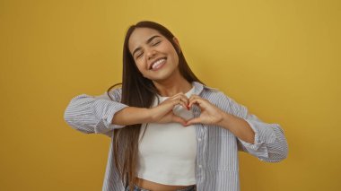 Kadın gülümseyen ve elleri ile bir kalp oluşturan canlı sarı bir arka plan, mutluluk ve sıcaklık ifade eden, gündelik bir gömlek ve uzun esmer saç.