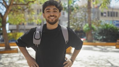 Sırt çantası takmış, şehir parkında gülümseyen sakallı yakışıklı bir Arap..