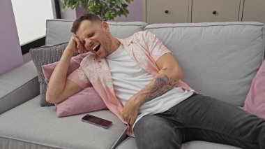 Modern bir oturma odasında, akıllı telefonuyla gri bir koltukta oturan, gülen dövmeli bir adam..