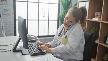 Genç sarışın bir kadın, bir doktor. Parlak bir hastane ofisinde telefon görüşmesi yaparken bilgisayarda birden fazla iş yapıyor.