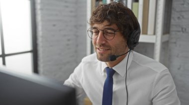 Modern ofis ortamında kulaklık takan genç adam bilgisayarla iletişim kurarken gülümsüyor ve profesyonel iş ortamını içeride gösteriyor..