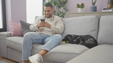 Genç bir İspanyol adam telefonuyla bir kanepede dachshund köpeğinin yanında dinleniyor, evde sıradan bir günü canlandırıyor..