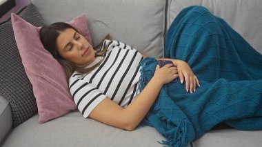 Mavi bir battaniyenin altında, rahat bir oturma odasında uyuyan esmer bir kadın.