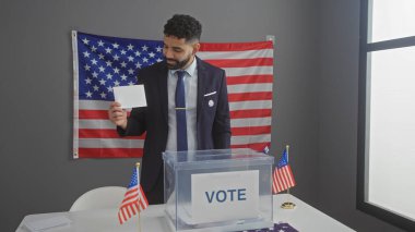 Genç bir İspanyol, arka planda bayrakları olan bir Amerikan seçim merkezinde oy pusulasını inceliyor..