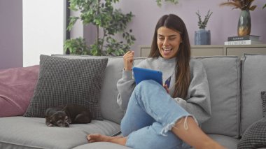 Gülümseyen genç kadın rahat oturma odasında uyuyan bir chihuahua ile kanepede tablet yemekten hoşlanıyor.