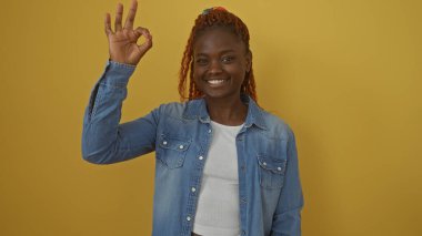 Kot giymiş gülümseyen genç siyah kadın sarı arka planda izole edilmiş iyi bir jest yapıyor.