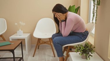 Çekici bir İspanyol esmer kadın, modern dekorlu ve köşesinde bir bitki olan, kapalı bir odada düşünceli bir şekilde oturur..