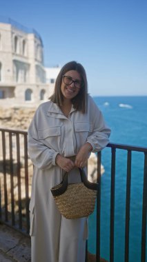 Polignano 'daki bakış açısının tadını çıkaran genç İspanyol kadın bir kısrak, puglia, İtalya, arka planda deniz ve tarihi mimariyle birlikte bir kamış torba tutarken gülümsüyor..