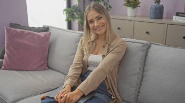 Genç, güzel, sarışın, beyaz bir kadın şık bir oturma odasında oturuyor, elinde dizüstü bilgisayarla evde rahat bir şekilde gülümsüyor..
