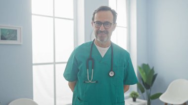 Önlüklü, steteskoplu, parlak bir hastane odasında duran kendinden emin erkek sağlık uzmanı..