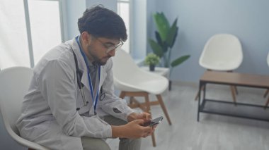Beyaz önlüklü yakışıklı Arap hastane bekleme odasında akıllı telefon kullanıyor..