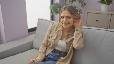 Genç, çekici, sarışın bir kadın oturma odasındaki gri bir kanepede oturuyor, elini kulağına uzatmış bir jest yapıyor..