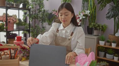 Genç Asyalı bir kadın iç mekandaki bir dükkanda çiçeklerini düzenliyor. Etrafı yapraklarla çevrili bir dizüstü bilgisayarla uğraşırken..