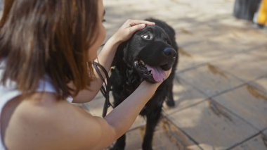 Güneşli bir günde siyah labrador köpeğini okşayan genç, güzel bir İspanyol kadın evcil hayvanıyla iç açıcı bir anı yakalıyor..
