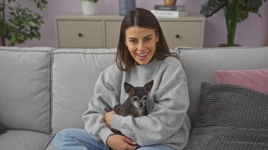 Smiling young woman holding a chihuahua in a cozy modern living room. clipart