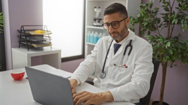 Klinikte dizüstü bilgisayar üzerinde çalışan, gözlük ve steteskop takan, bitkilerle çevrili, profesyonel bir hastane ortamında iş yerindeki görevlere odaklanan bir doktor..