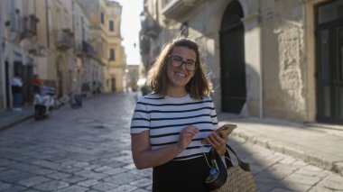 İtalya, Lecce 'de genç bir İspanyol kadın elinde akıllı bir telefon, büyüleyici tarihi mimarisi ile güneşli bir şehir sokağında dururken gülümsüyor..