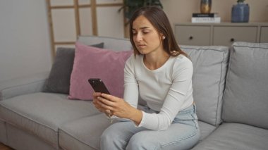 Genç, beyaz, çekici bir kadın evinde rahat bir oturma odasında kanepede otururken akıllı telefon kullanıyor..