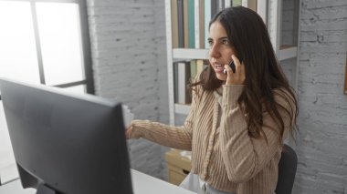 Modern ofiste telefonla konuşan bir kadın bilgisayar, esmer ve çekici, profesyonel iş yerinde kitaplık ile kazak giyiyor, verimliliği ve iletişimi resmediyor.