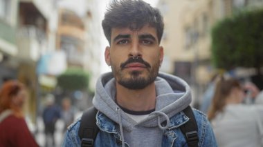 Günlük kıyafetleriyle sakallı yakışıklı İspanyol bir adam işlek bir caddede dikiliyor, düşünceli görünüyor..