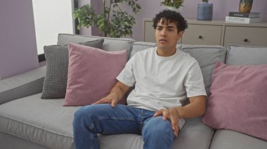 Modern bir oturma odasında gri bir koltukta uzanan genç bir İspanyol adam dekoratif yastıklar ve ev bitkileriyle iç içe.