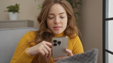 Sarı kazak giyen genç güzel sarışın kadın rahat oturma odasında oturuyor ve rahat bir ifadeyle akıllı telefonunu kullanıyor. Etrafı kapalı bitkiler ve yumuşak mobilyalarla çevrili..