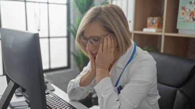 Klinikteki yaşlı bir kadın masada otururken stresli görünüyor ve bilgisayar kullanıyor, dalgın ve yorgun görünüyor..