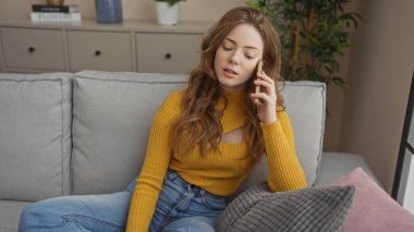 Sarışın, çekici bir genç kadın oturma odasında bir kanepede oturuyor, telefonla konuşuyor, sarı bir kazak ve kot pantolon giyiyor, bir apartmanda, rahat görünüyor..