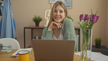 Ev ofis masasında gözlüklü, dizüstü bilgisayarı, kulaklığı ve rahat bir odada laleleri olan gülümseyen genç bir kadın..