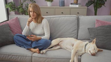 Akıllı telefonlu düşünceli bir kadın rahat bir oturma odasında uyuyan köpeğin yanında koltukta oturuyor.