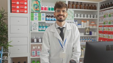 Sakallı, laboratuvar önlüğü giymiş, eczanenin içinde duran gülümseyen İspanyol genç adam..