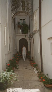 Güzel, genç bir İspanyol kadın Locorotondo, Puglia, İtalya 'da dar bir sokakta yürüyor. Etrafı beyaz binalar ve canlı saksı bitkileriyle çevrili..