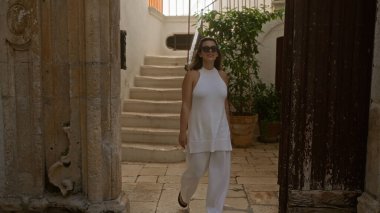 Güzel, genç, İspanyol bir kadın, büyüleyici Avrupa mimarisine karşı beyaz giyinmiş, Locorotondo, Puglia, İtalya 'nın eski sokaklarında yürüyor..