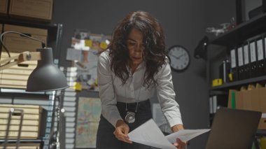 Esmer bir kadın dedektif, soruşturmayı ve kolluk kuvvetlerini sembolize eden loş bir ofis odasında belgeleri inceliyor..