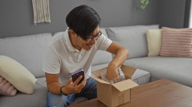 Genç adam paketini baloncuklu ambalajla açıyor. Elinde akıllı bir telefon, oturma odasında kanepede oturuyor.