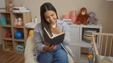 Yatak odasında beşiği ve oyuncaklarıyla kitap okuyan, peluş hayvanlar ve bebek eşyalarıyla çevrili, gülümseyen ve huzurlu çocuk odası ortamının tadını çıkaran bir kadın..