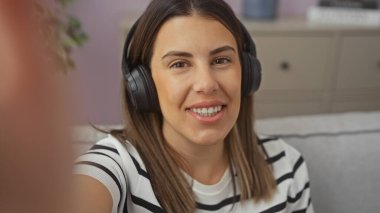 Beautiful young hispanic woman taking a selfie at home in the living room, wearing headphones and a striped shirt, sitting on a couch in a relaxed indoor setting. clipart