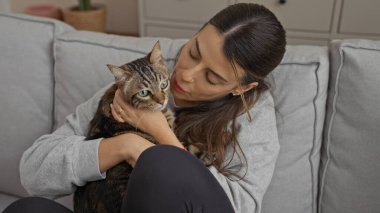 Evinin oturma odasında evcil kedisine sarılan genç bir İspanyol kadın..